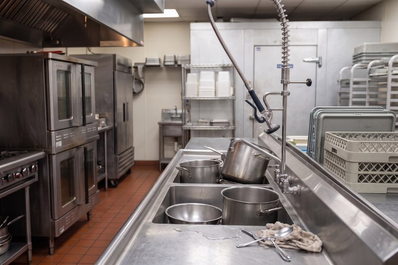 Professional plumber with the Affordable Plumbing logo cap installing a pre-rinse faucet in a commercial kitchen sink.